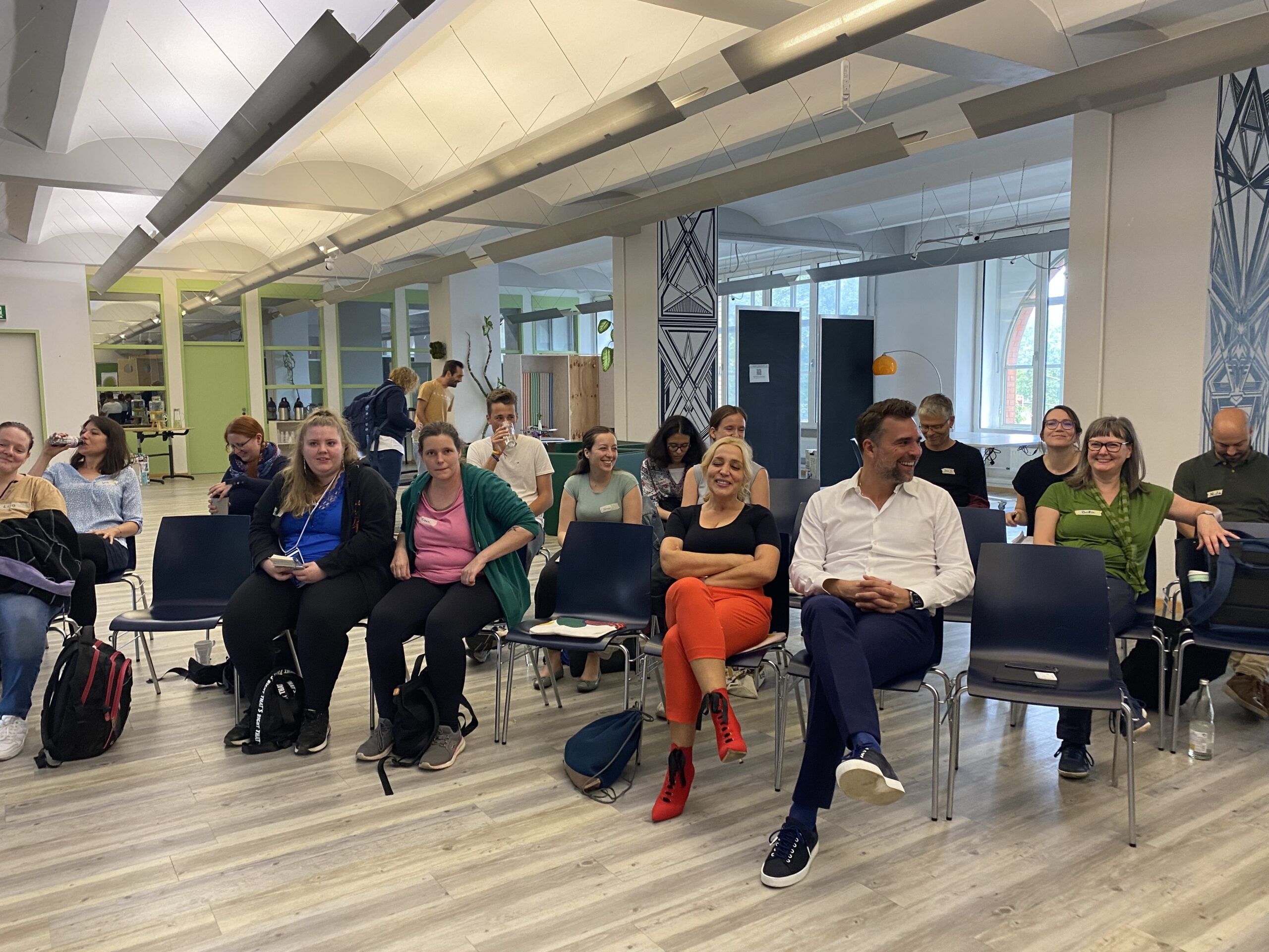 Teilnehmende sitzen auf Stühlen verteilt und sind alle nach vorne ausgerichtet. Ein Lachen geht durch die Runde.