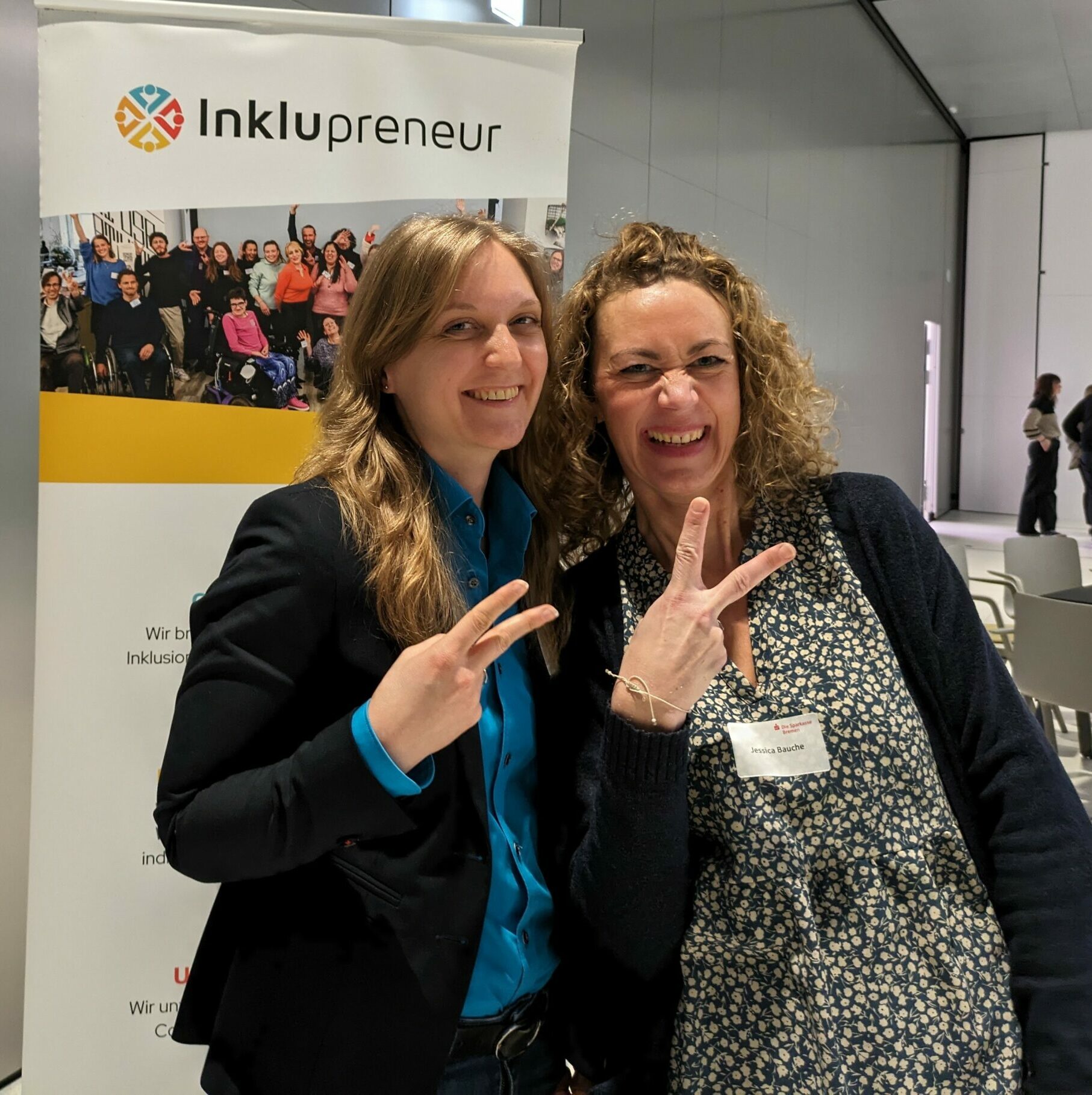 Zwei Frauen stehen vor einem Aufsteller von Inklupreneur und lachen.